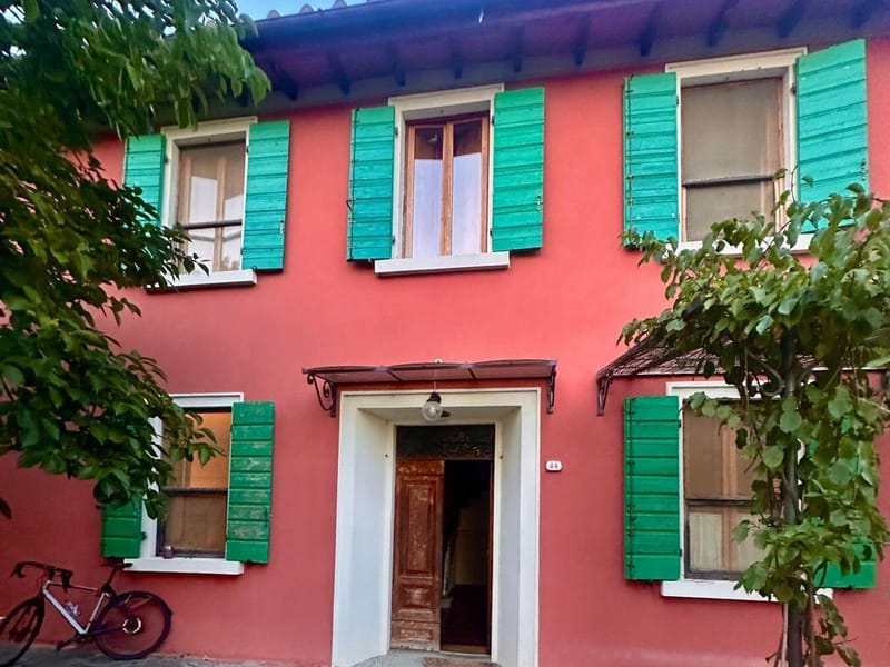 A pink Tuscan farmhouse with green shutters, a stone courtyard, and a bicycle leaning against the wall