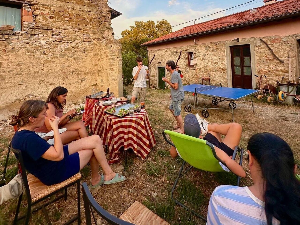 A group of coliving guests eating and relaxing at an outdoor table beside a stone farmhouse in Tuscany, with a ping pong table and wooded hills in the background