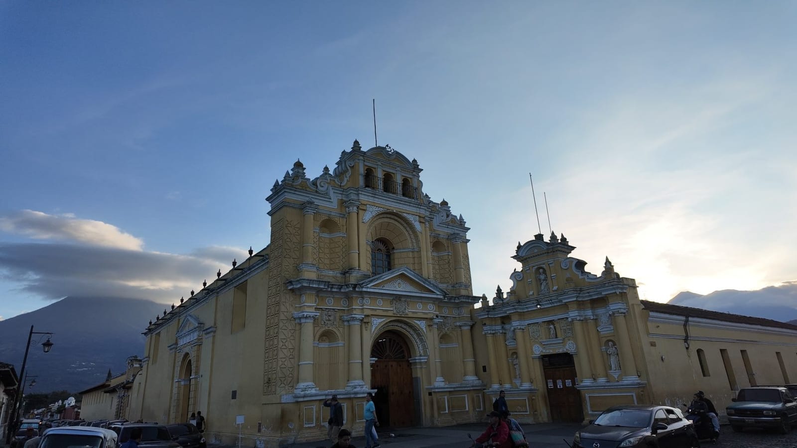 The ochre Baroque facade of a colonial-era church in Antigua Guatemala, lit by early evening sky with a volcano silhouette rising behind the rooftops and a cobblestone street busy with people and cars in front