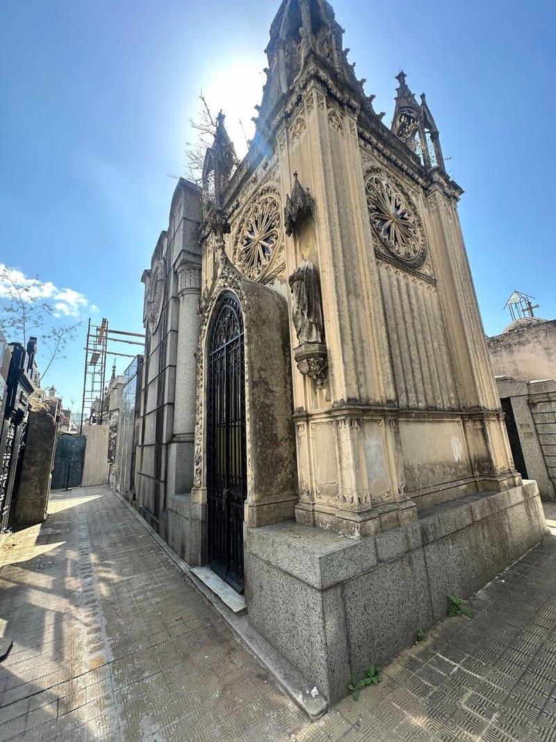 A tall Gothic-style stone chapel tower with rose windows and carved spires, seen from a cemetery walkway under a clear blue sky