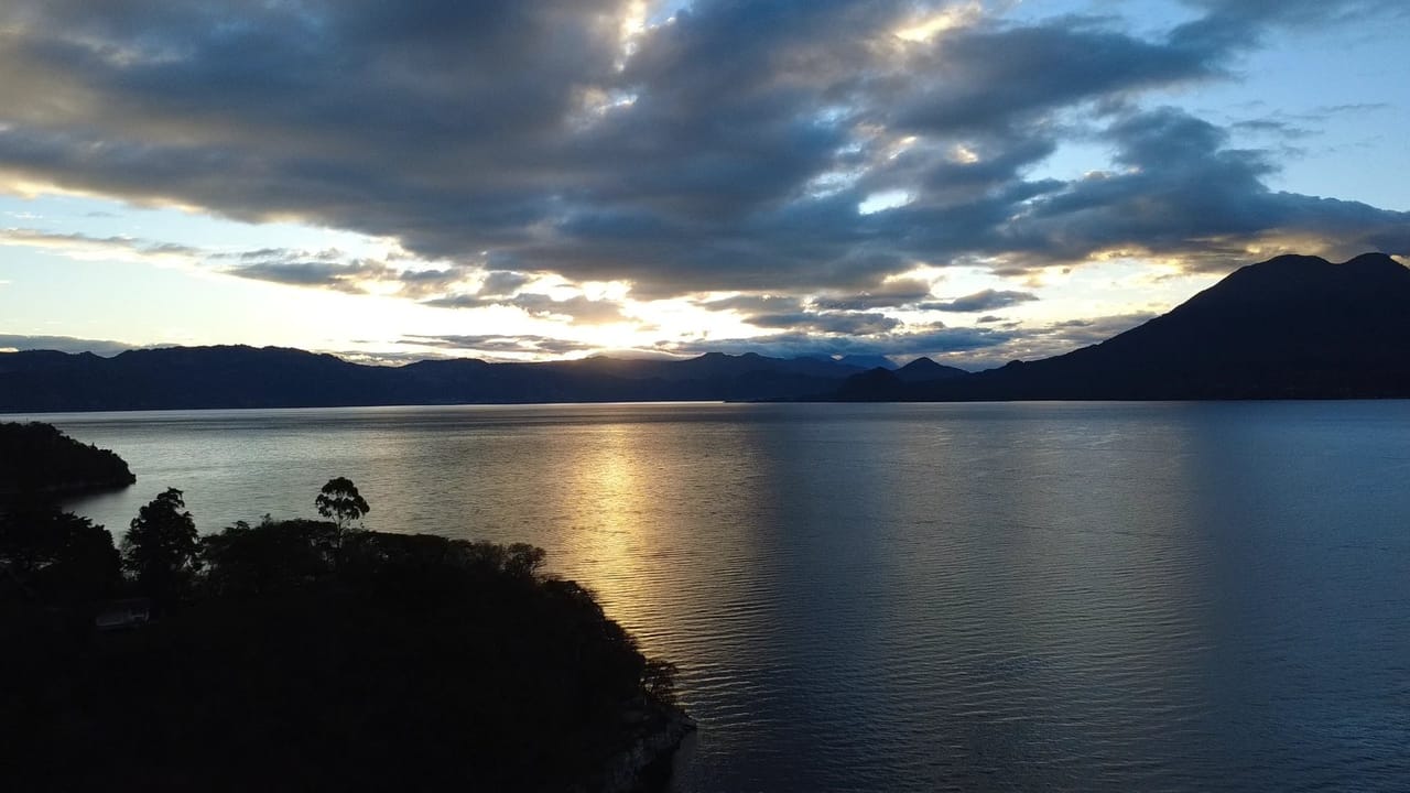 Sunrise breaking through storm clouds over Lake Atitlán, with volcano silhouettes across the water and a forested point in the foreground