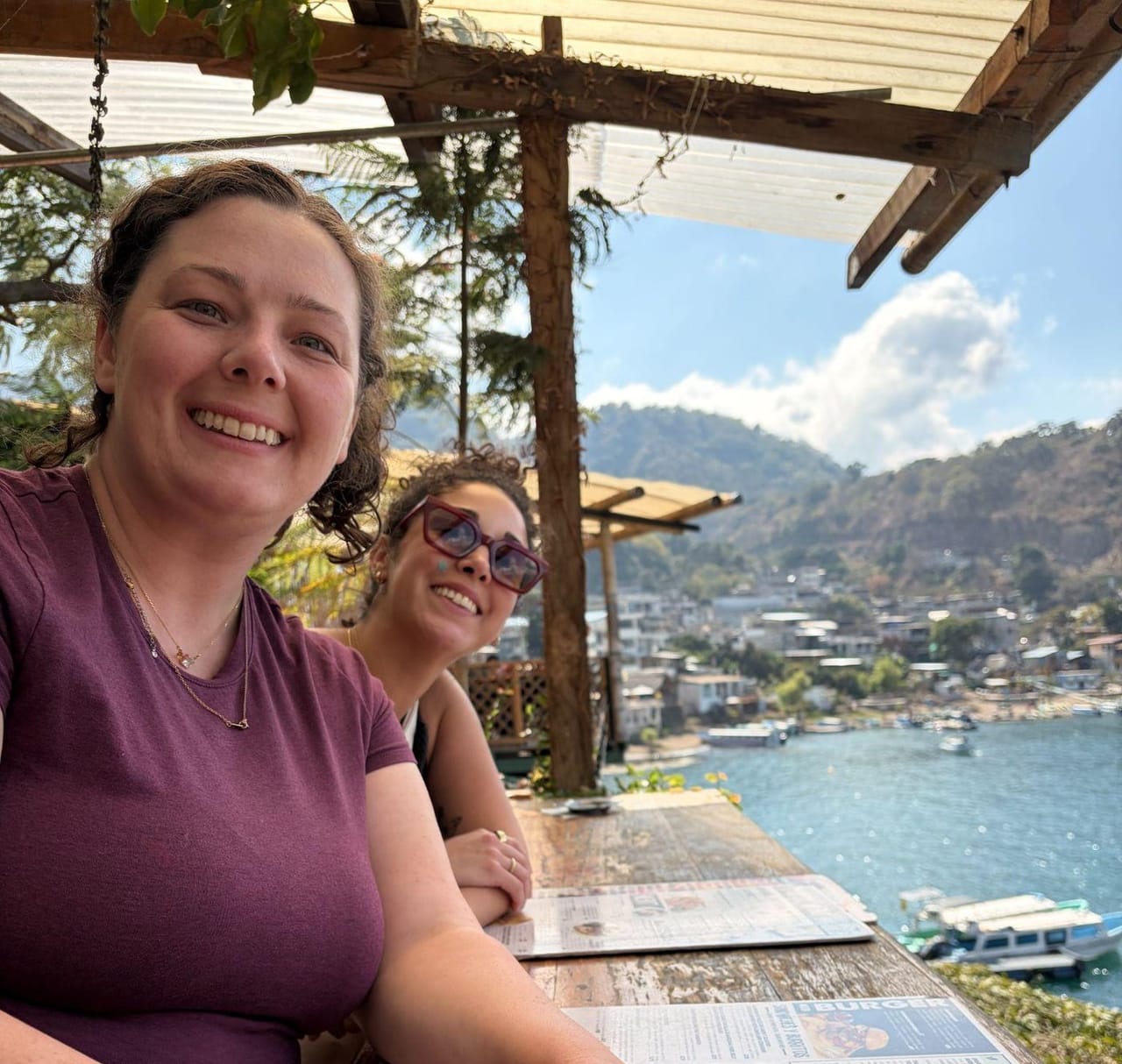 Two women smiling at an outdoor table at Bar Sublime, with menus in front of them and a view of Lake Atitlán, boats, and mountains behind them