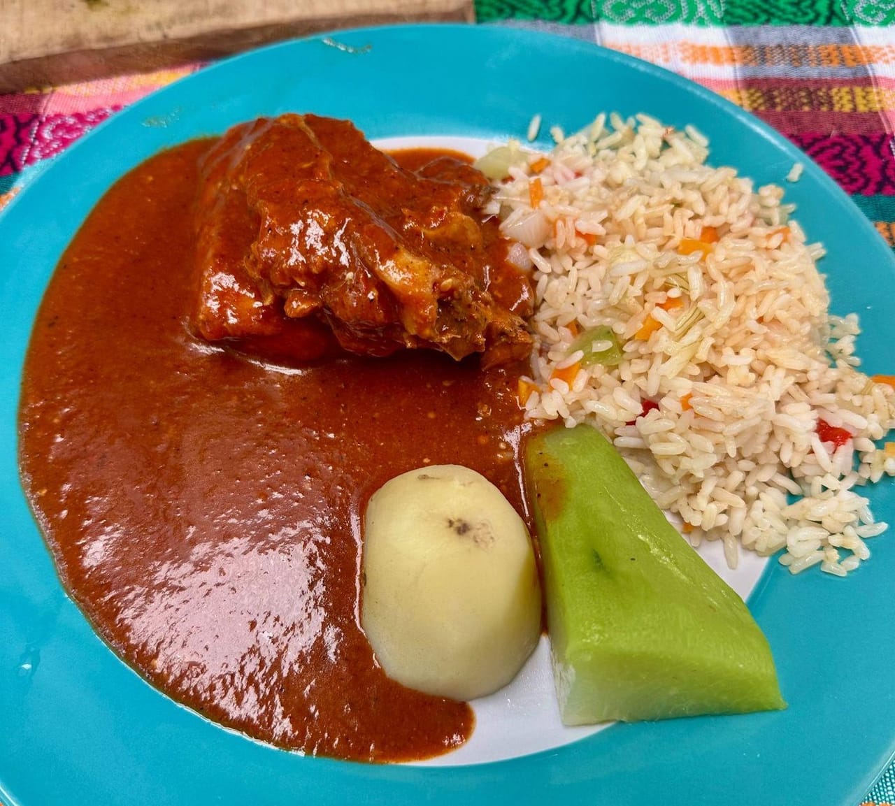 A plate of pepián stew with a piece of meat in red sauce, rice, a boiled potato, and a wedge of chayote, served on a teal plate over a woven textile