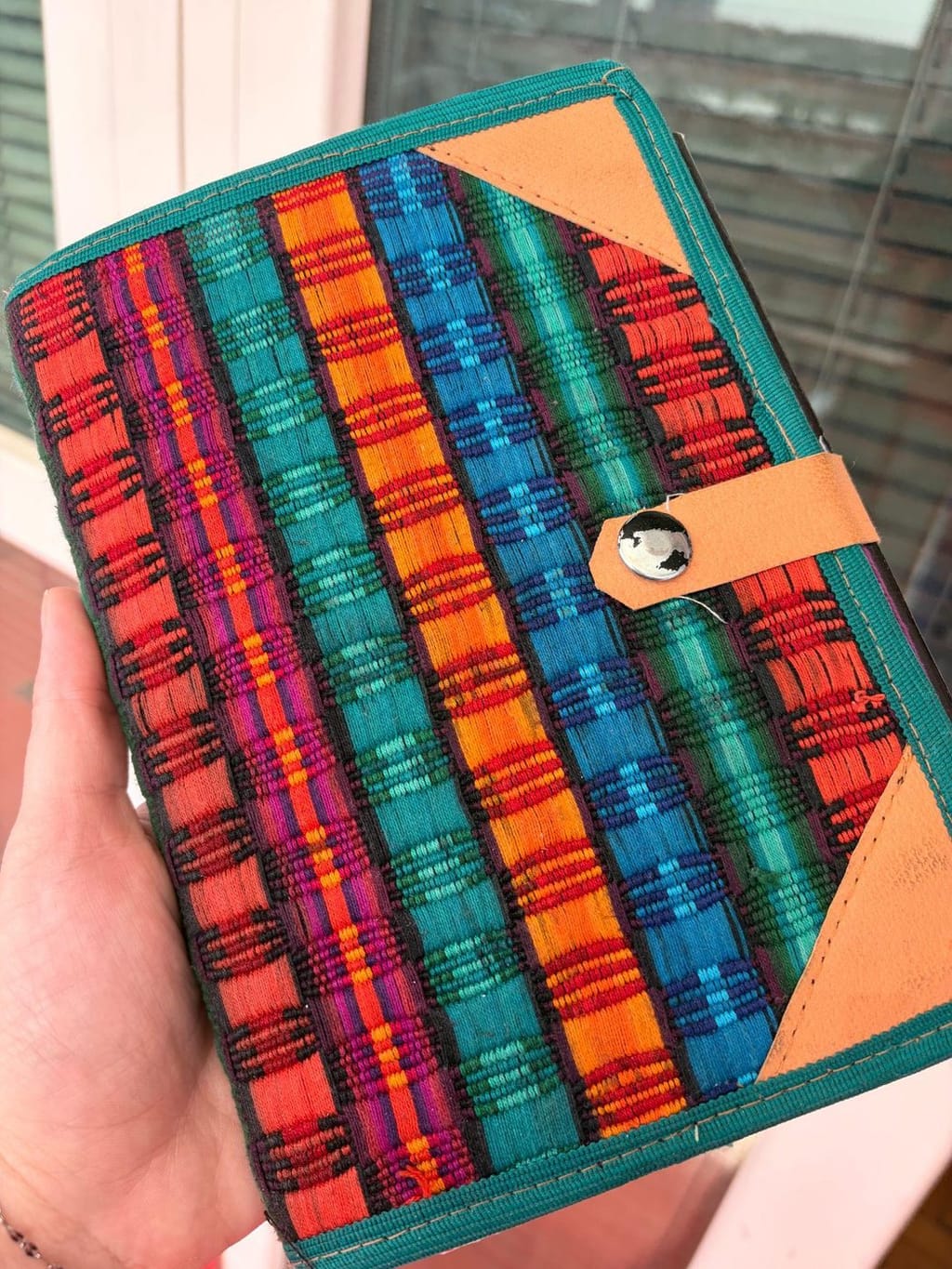 A hand holding a woven fabric journal cover in red, teal, orange, and blue stripes, with a tan leather snap closure