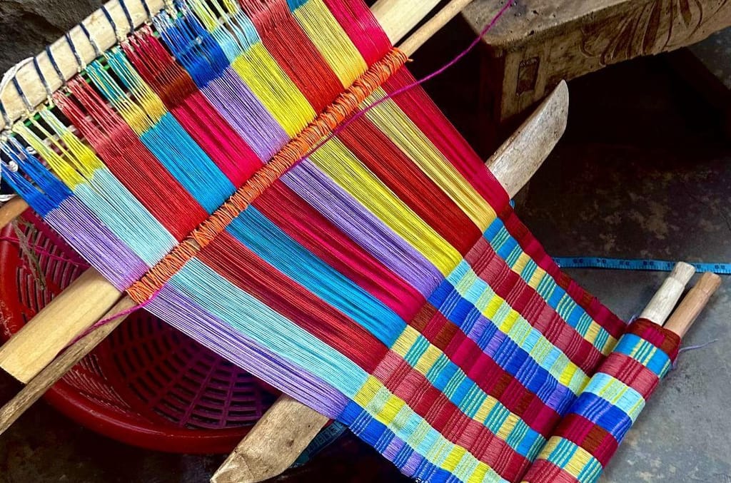 A partially finished backstrap weaving in progress, with brightly colored red, yellow, blue, and purple threads strung on a wooden loom