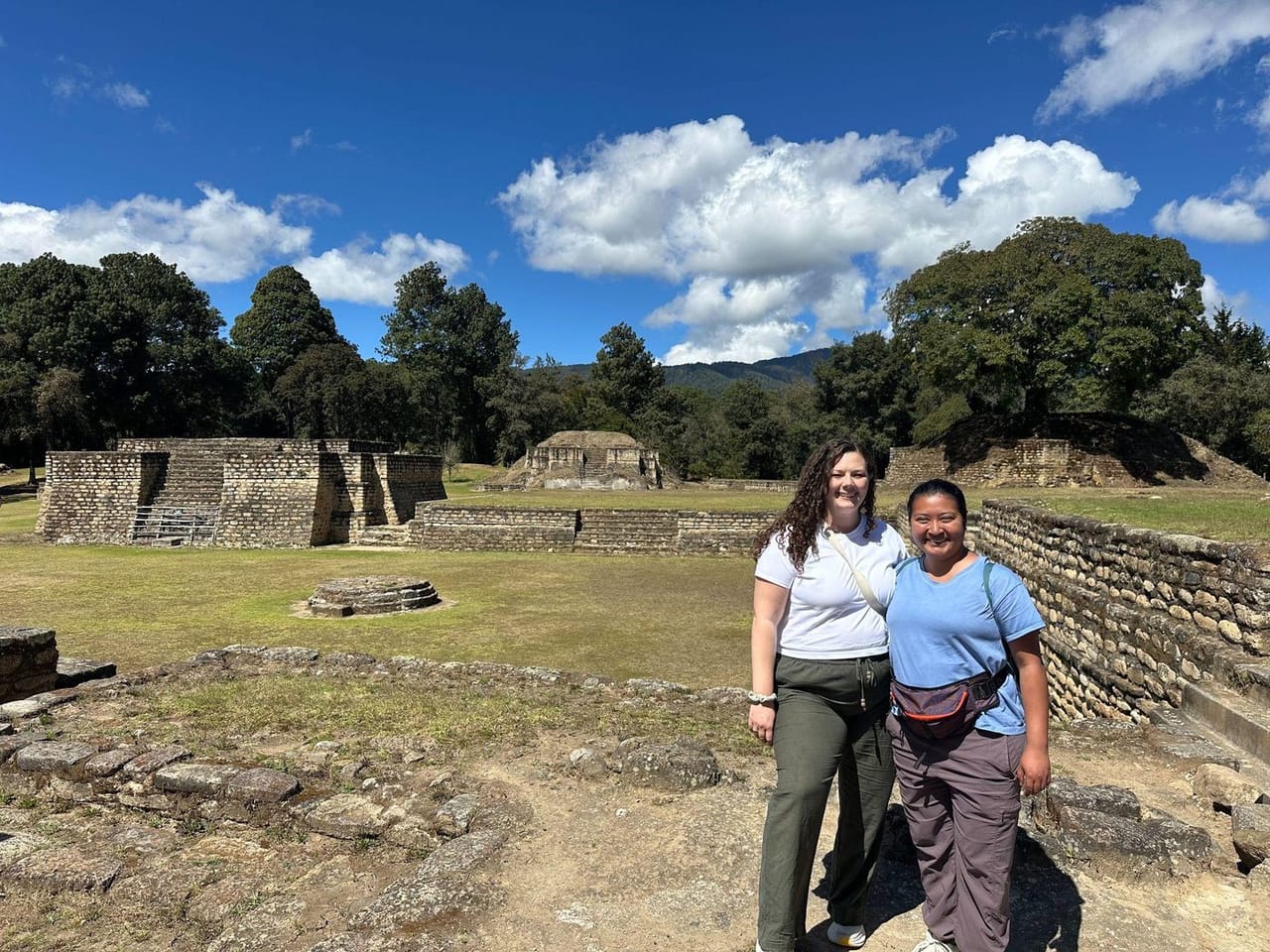 Two travelers standing among the low, weathered stone ruins at Iximche
