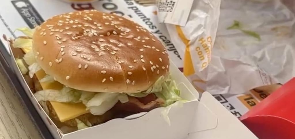Close-up of a sesame-seed-bun chicken sandwich with cheese, lettuce, and bacon in an open cardboard box on a McDonald's tray