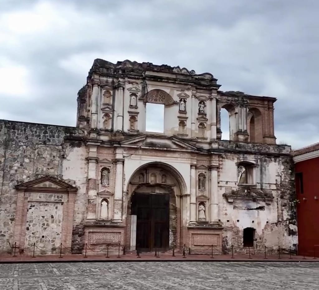 The roofless, weathered stone facade of a ruined colonial church in Antigua Guatemala, with carved columns and empty window openings under a cloudy sky