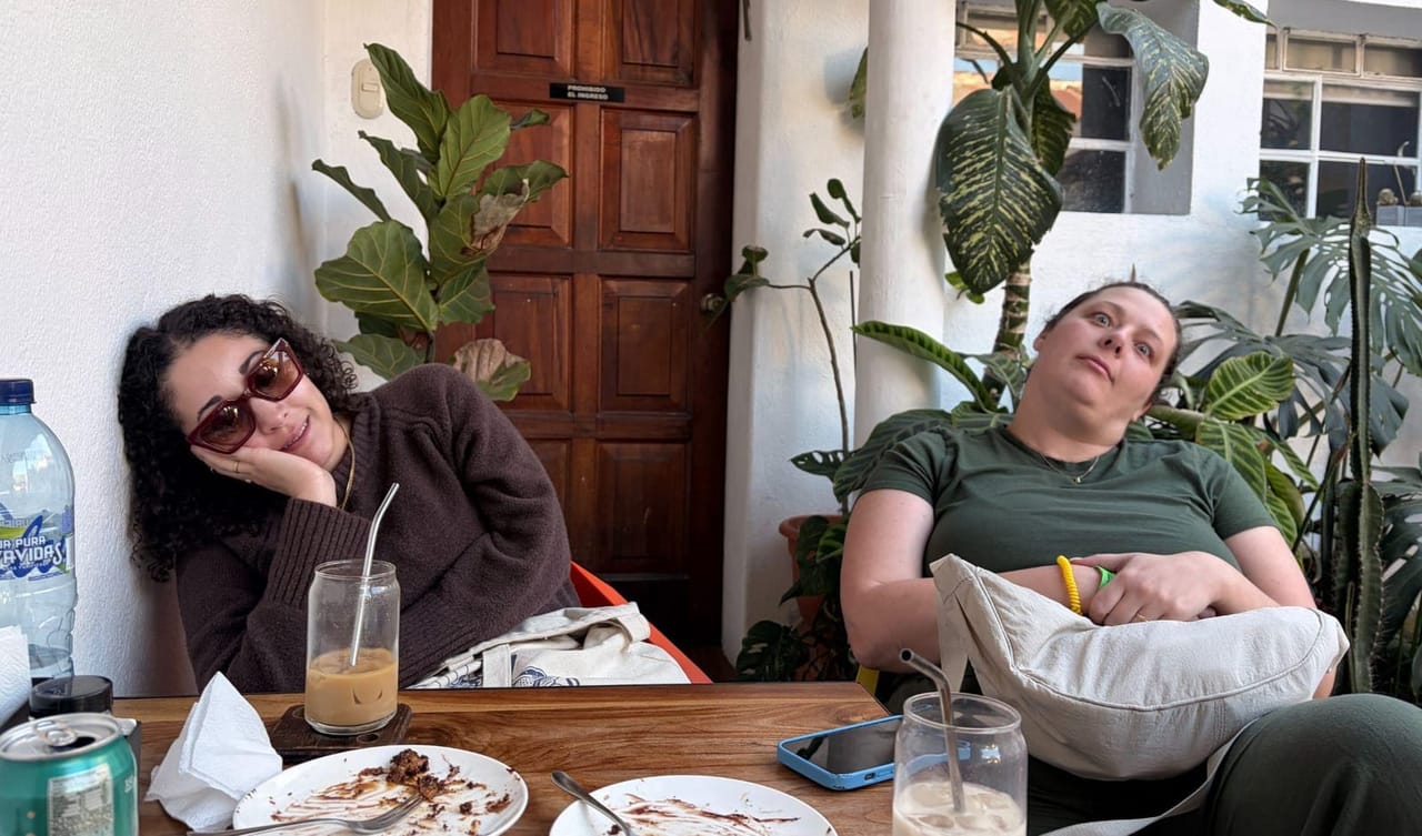 Two women slumped contentedly in their chairs at a cafe table with empty plates and iced drinks, in a plant-filled courtyard