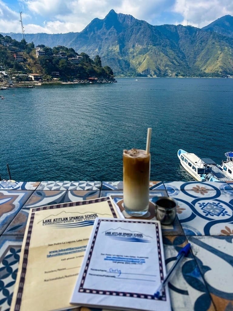 Iced coffee, a pen, and two Lake Atitlan Spanish School booklets on a patterned tile table overlooking the lake, with a passenger boat docked below and steep green mountains across the water