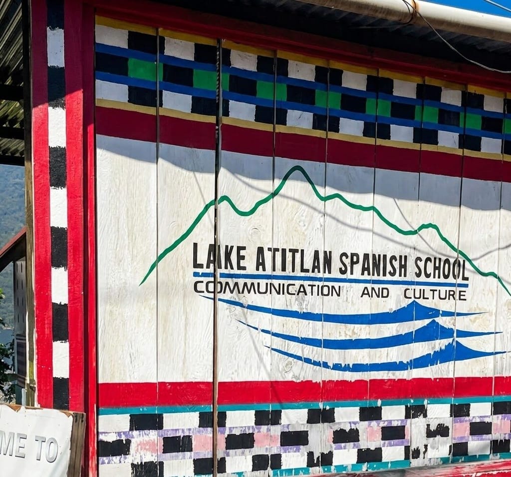 Hand-painted sign on a wooden wall reading “Lake Atitlan Spanish School, Communication and Culture,” with a green mountain outline, blue waves, and red, black, and white patterned borders