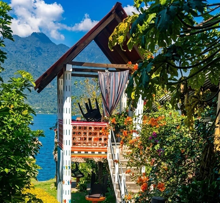 Open-air classroom on the upper level of a peaked-roof hut, reached by outdoor stairs lined with orange flowers, with the lake and a forested mountain behind