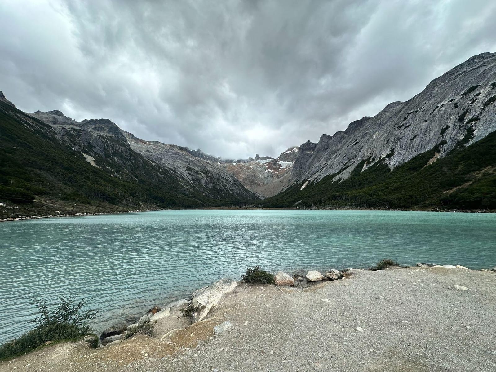 A turquoise glacial lake ringed by a forested mountain valley under a cloudy sky, with a rocky shoreline in the foreground