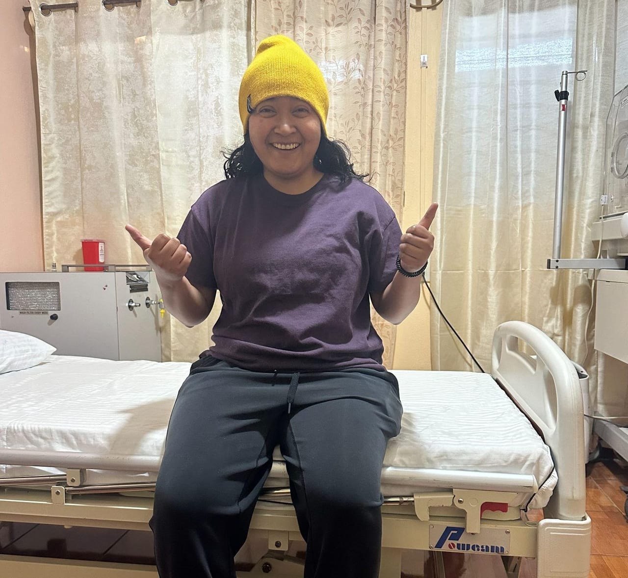 Jackie lying in a hospital bed hooked up to an IV drip at a clinic near Lake Atitlán, giving a thumbs up