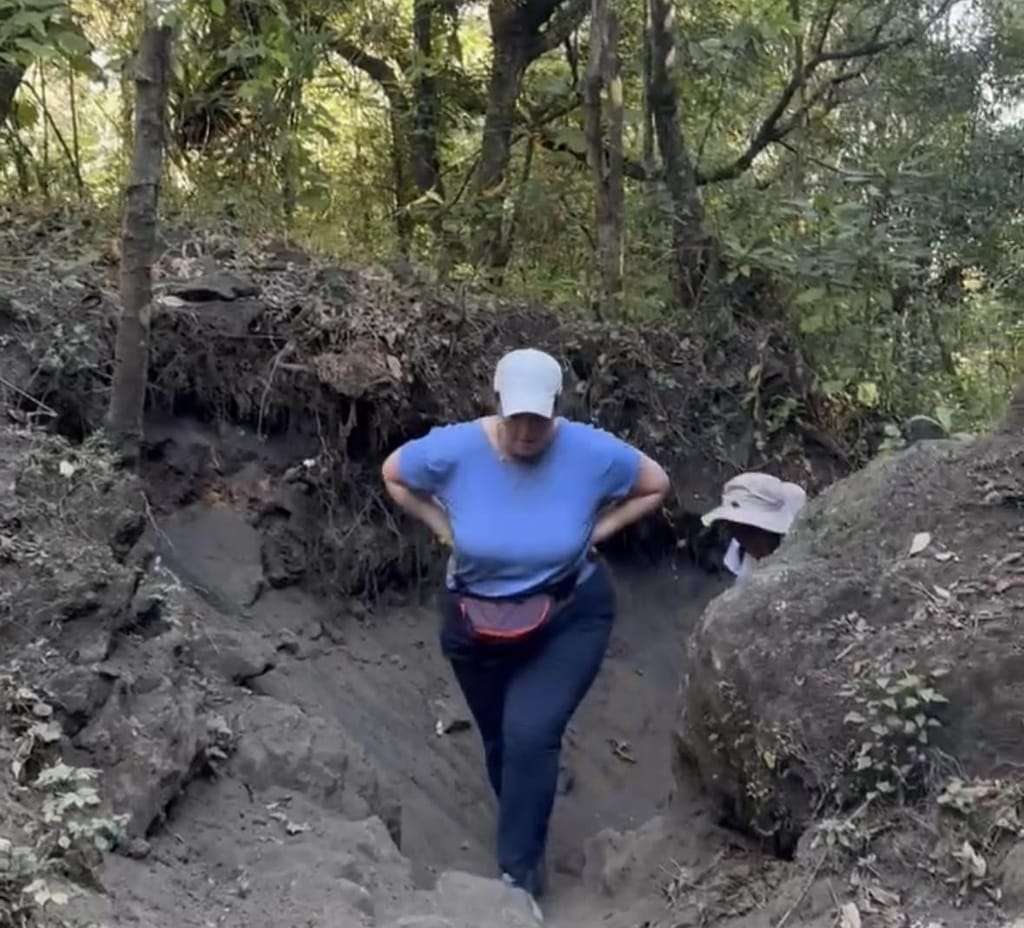Carly, in a white cap and blue T-shirt, bending forward with her hands on her hips as she climbs a steep, eroded dirt trail through the forest