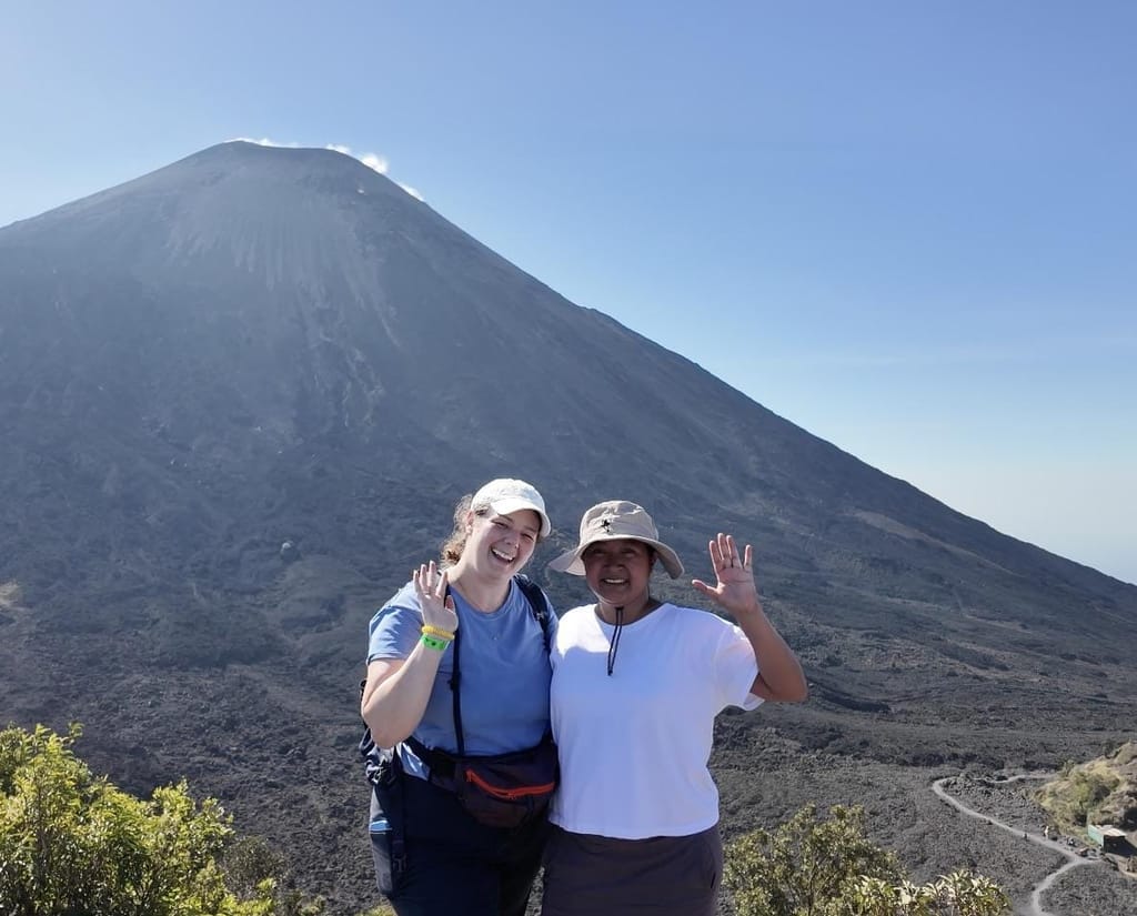 Carly and Jackie smiling with their arms around each other, Jackie waving, in front of the dark, bare cone of Volcán Pacaya