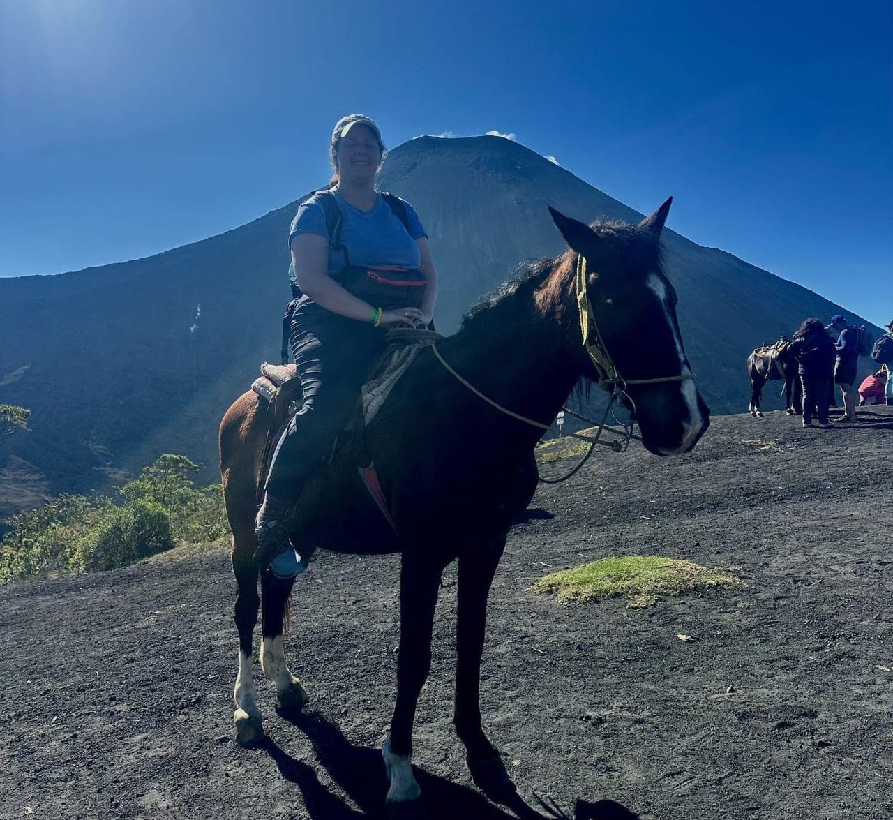 Carly sitting on a dark brown horse on the black volcanic sand of Pacaya's plateau, with the volcano's cone rising behind her against a deep blue sky