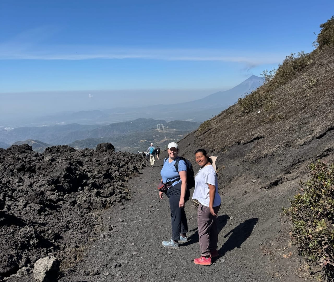 Jackie and Carly standing together on a dark volcanic gravel trail on Pacaya, with a steep rocky slope beside them and hazy hills and valleys stretching into the distance