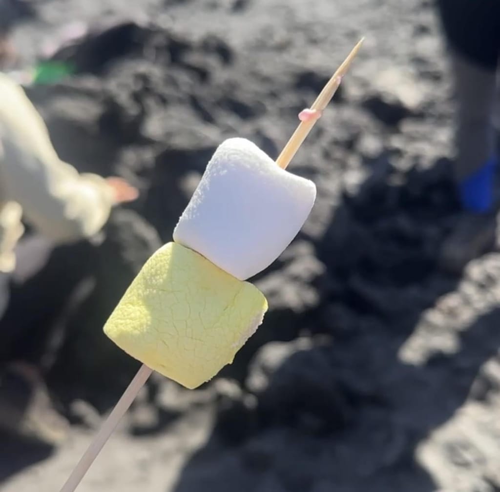 Close-up of two marshmallows on a wooden skewer, the lower one toasted yellow, held above dark volcanic ground