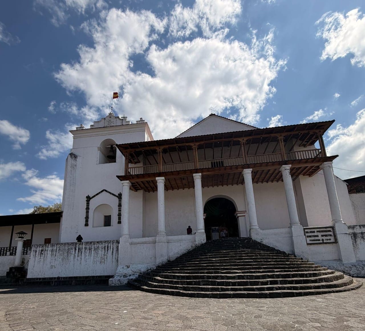 White colonial Church of Santiago Apóstol in Santiago Atitlán, Guatemala, with a bell tower, wooden balcony, white columns and curved stone steps leading up from a cobblestone plaza under a blue sky. A sign beside the entrance reads “Iglesia Parroquial Santiago Apóstol, Fundada 1547.”