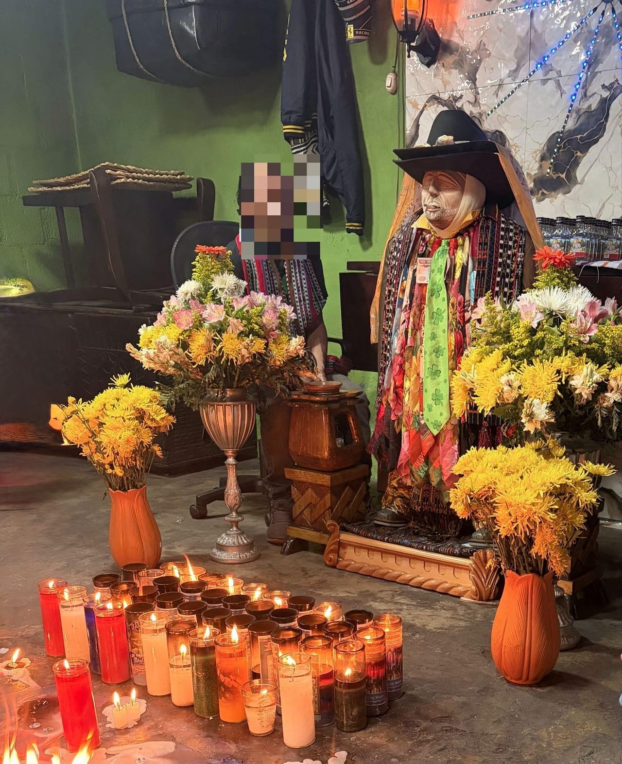 Maximón shrine in Santiago Atitlán, Guatemala. The wooden effigy wears a black cowboy hat, a cigarette at its lips, and layers of colorful silk ties and scarves. It stands among large arrangements of yellow, white and pink flowers, with rows of lit prayer candles on the floor in front.