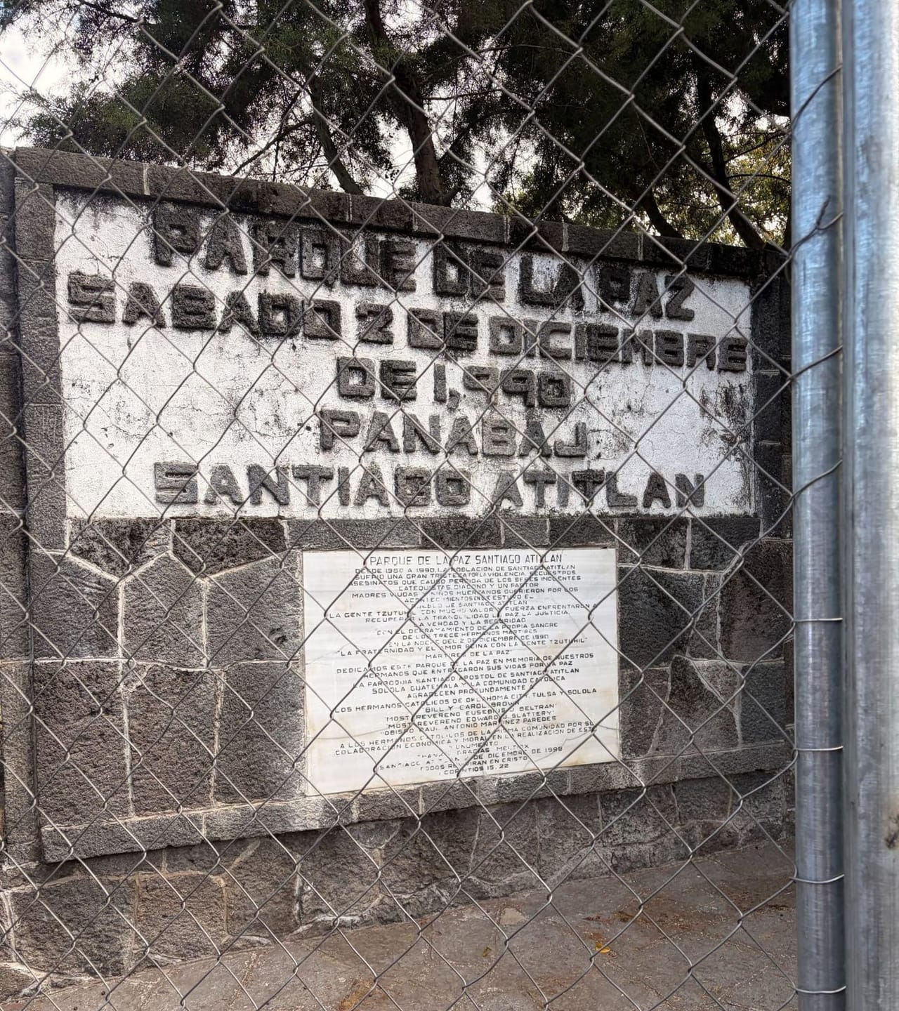 Stone wall seen through a chain-link fence, with raised lettering reading “Parque de la Paz, Sábado 2 de Diciembre de 1990, Panabaj, Santiago Atitlán.” Below it, a white marble plaque honors the Tz’utujil residents killed in the 1990 massacre.