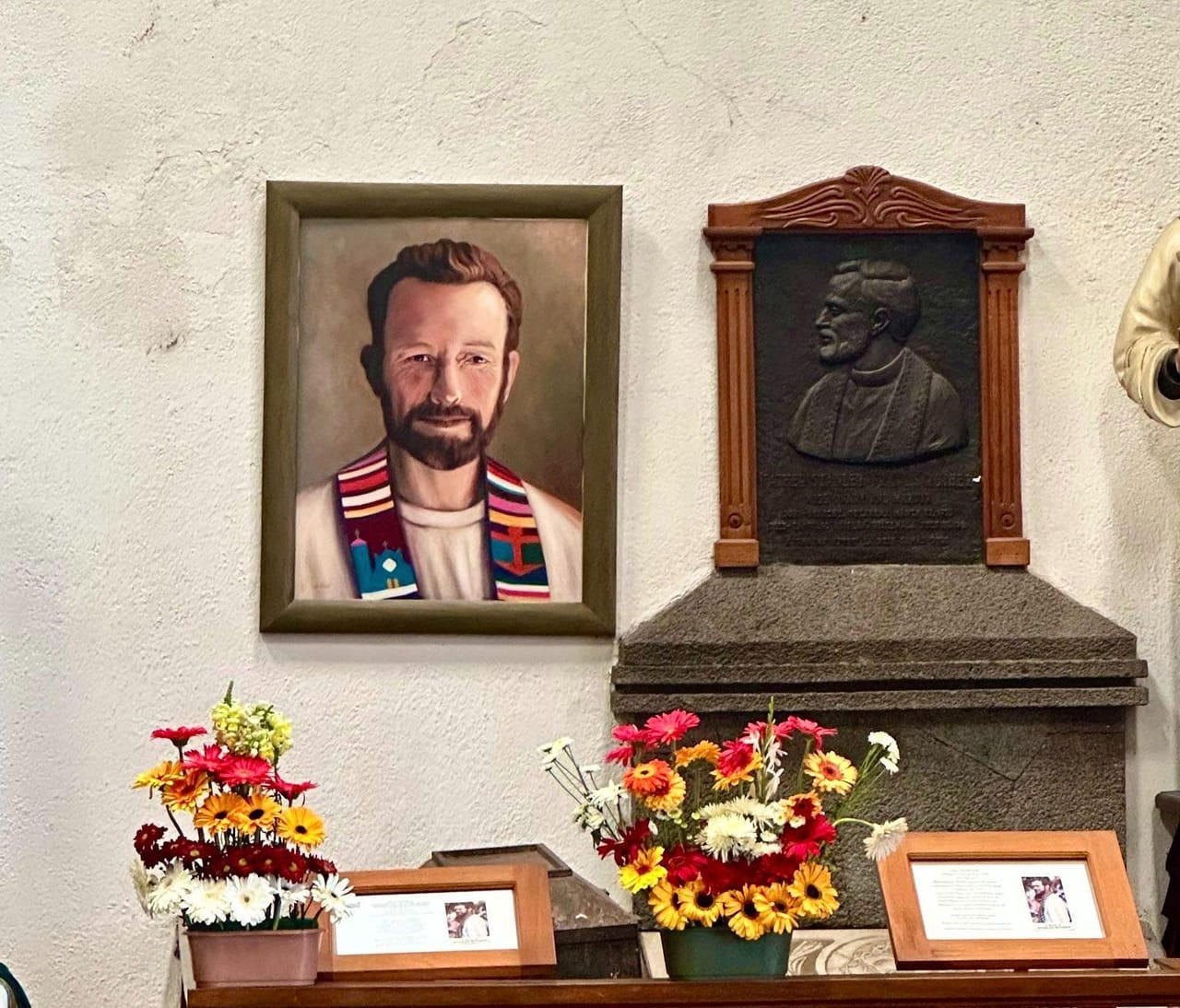A framed portrait of Father Stanley Rother beside a bronze memorial plaque, surrounded by flowers, inside a church in Santiago Atitlán